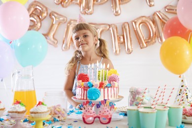Photo of Birthday celebration. Cute little girl at table with tasty cake and different treats in festive decorated room