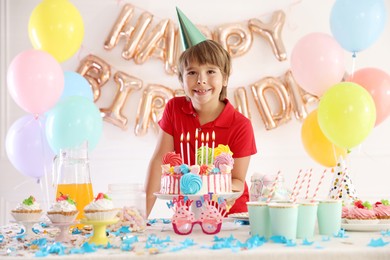 Photo of Birthday celebration. Cute little boy at table with tasty cake and different treats in festive decorated room