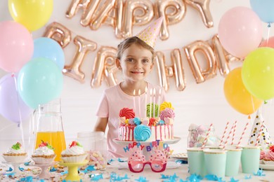 Photo of Birthday celebration. Cute little girl at table with tasty cake and different treats in festive decorated room