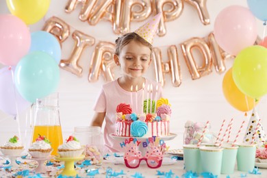 Photo of Birthday celebration. Cute little girl at table with tasty cake and different treats in festive decorated room