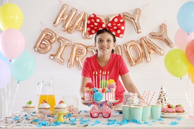 Photo of Birthday celebration. Cute girl at table with tasty cake and different treats in festive decorated room