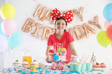 Photo of Birthday celebration. Cute girl at table with tasty cake and different treats in festive decorated room