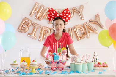 Photo of Birthday celebration. Cute little girl blowing out candles on cake and different treats in festive decorated room