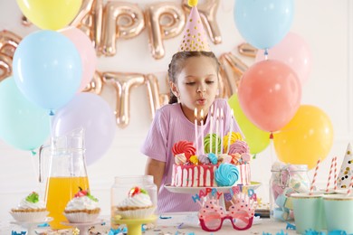 Photo of Birthday celebration. Cute little girl blowing out candles on cake and different treats in festive decorated room