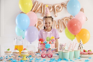 Photo of Birthday celebration. Cute little girl at table with tasty cake and different treats in festive decorated room