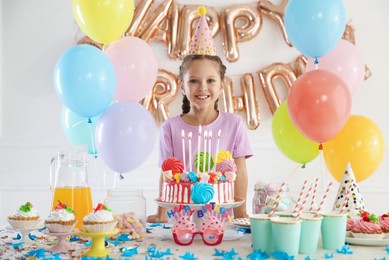 Photo of Birthday celebration. Cute little girl at table with tasty cake and different treats in festive decorated room