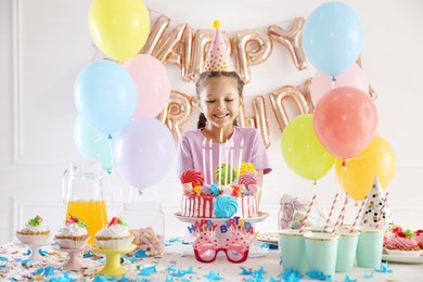 Photo of Birthday celebration. Cute little girl at table with tasty cake and different treats in festive decorated room