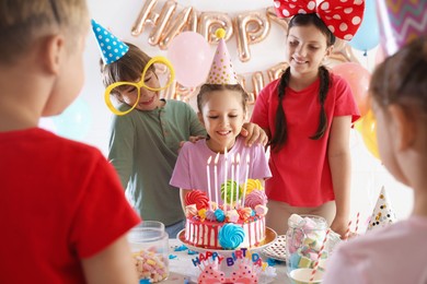 Photo of Birthday celebration. Cute little girl blowing out candles on cake and her friends in festive decorated room