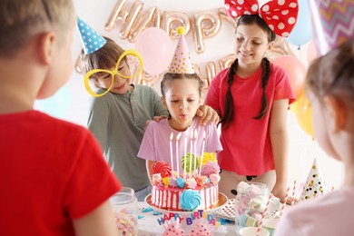 Photo of Birthday celebration. Cute little girl blowing out candles on cake and her friends in festive decorated room
