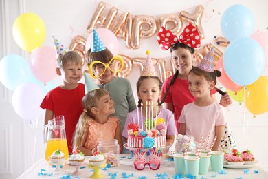 Photo of Birthday celebration. Cute little girl blowing out candles on cake at table and her friends in festive decorated room
