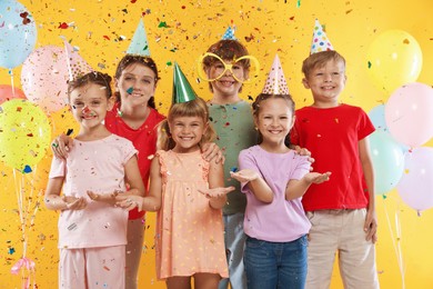 Photo of Birthday celebration. Happy kids in party hats under falling confetti on orange background