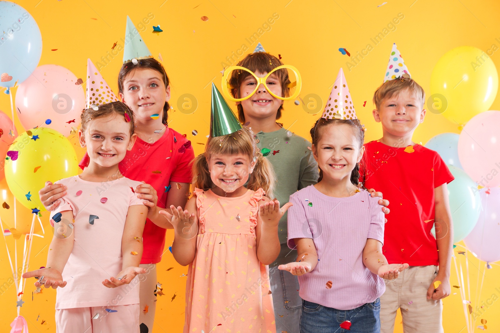 Birthday celebration. Happy kids in party hats under falling confetti on orange background Photo of Birthday celebration. Happy kids in party hats under falling confetti on orange background