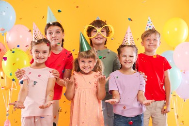 Photo of Birthday celebration. Happy kids in party hats under falling confetti on orange background