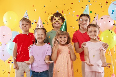 Photo of Birthday celebration. Happy kids in party hats under falling confetti on orange background