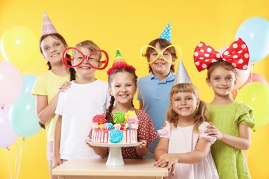 Photo of Birthday celebration. Happy kids with tasty cake and festive decor on yellow background