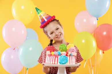 Photo of Birthday celebration. Cute little girl with tasty and balloons on orange background