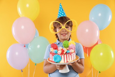 Photo of Birthday celebration. Cute little boy with tasty and balloons on orange background