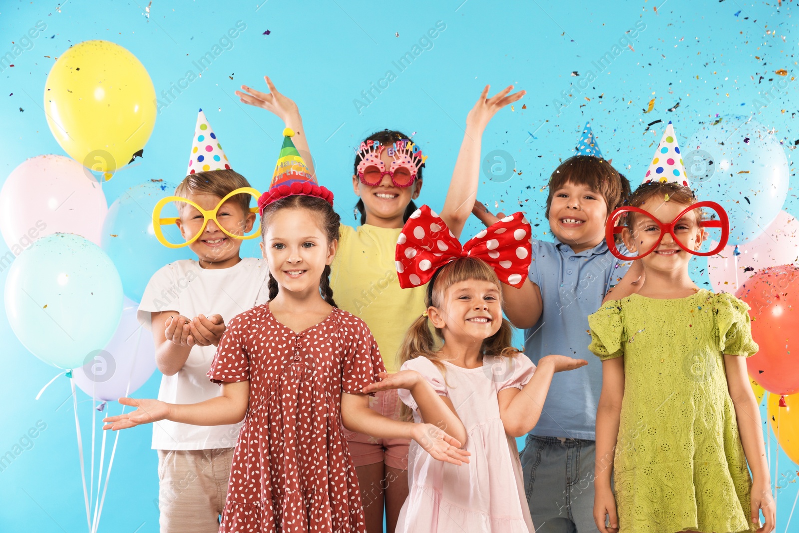 Birthday celebration. Happy kids with festive decor under falling confetti on light blue background Photo of Birthday celebration. Happy kids with festive decor under falling confetti on light blue background