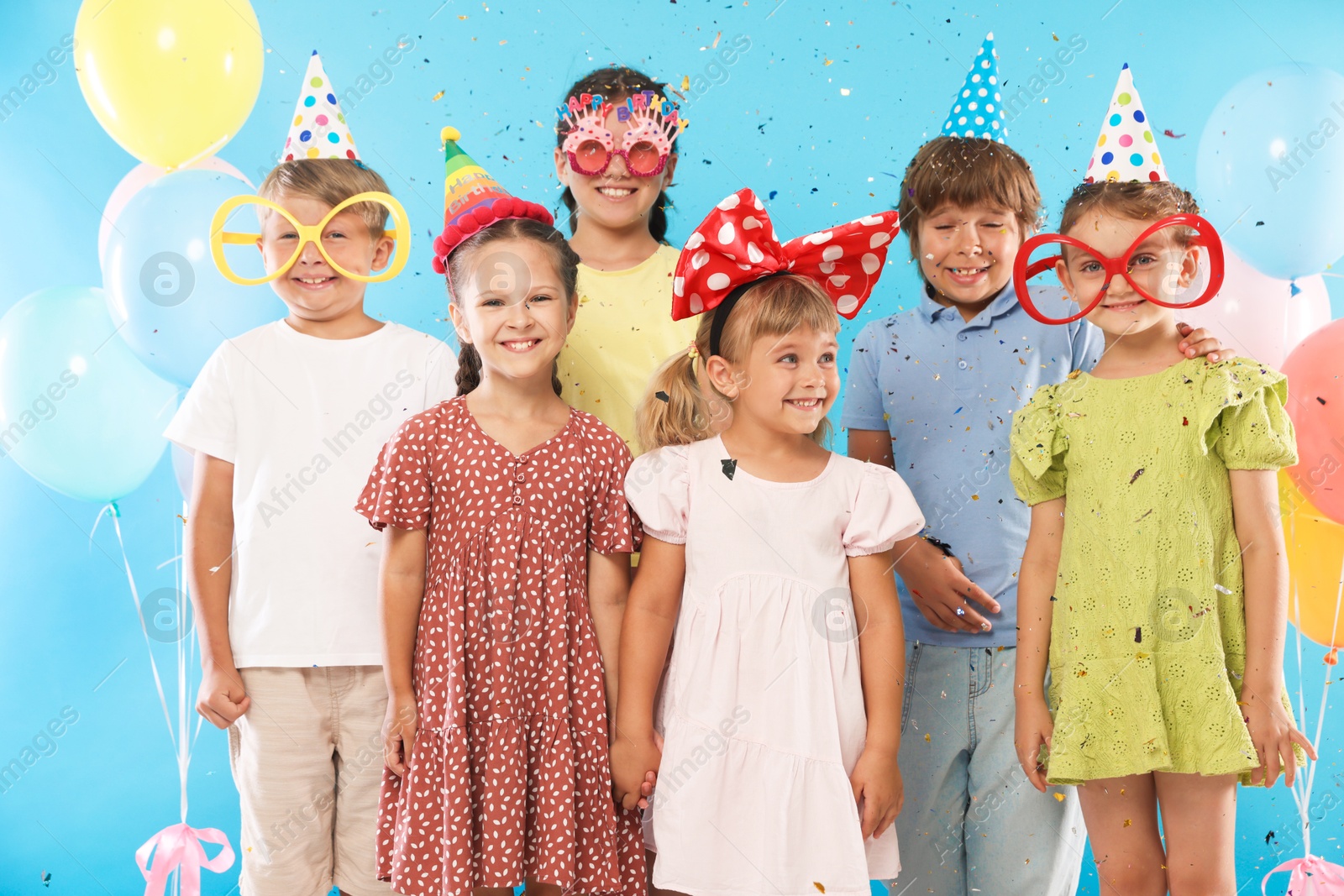Birthday celebration. Happy kids with festive decor under falling confetti on light blue background Photo of Birthday celebration. Happy kids with festive decor under falling confetti on light blue background