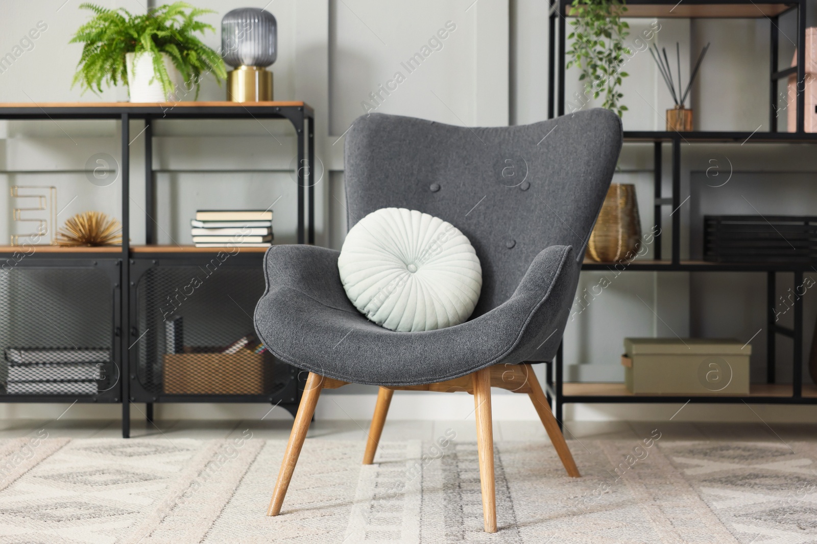 Photo of Stylish grey armchair with pillow and shelving unit indoors. Interior design