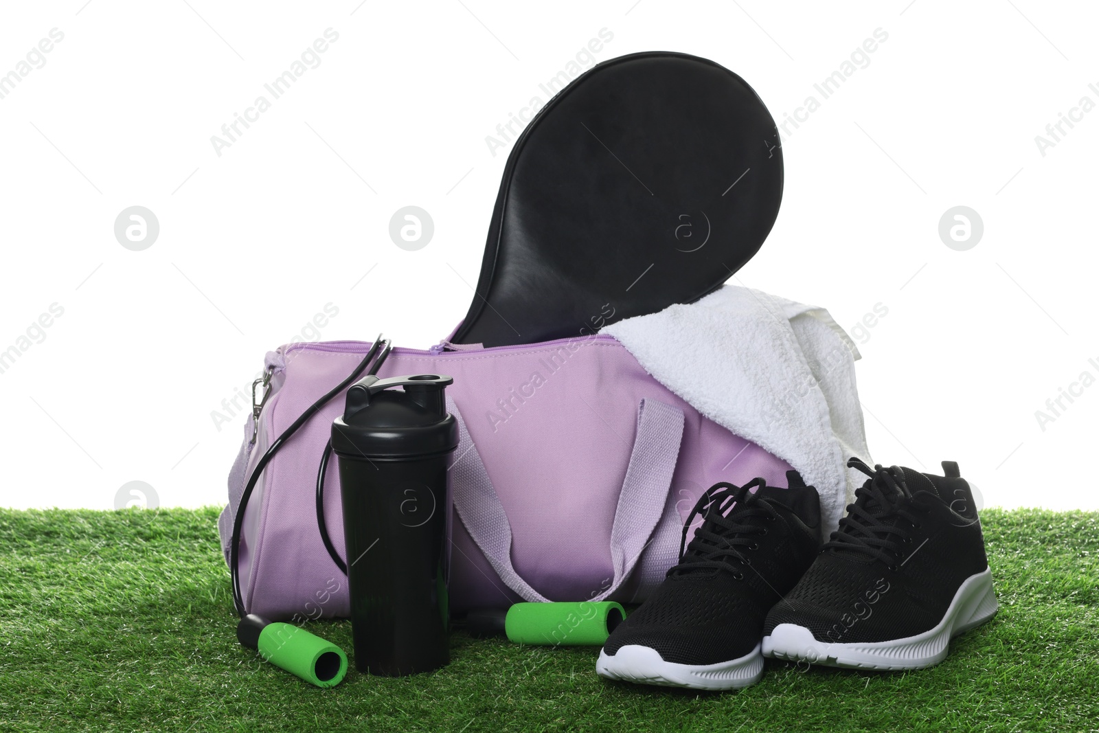 Photo of Stylish gym bag with sports equipment, towel, shaker cup and sneakers on green grass against white background