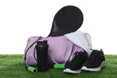 Photo of Stylish gym bag with sports equipment, towel, shaker cup and sneakers on green grass against white background