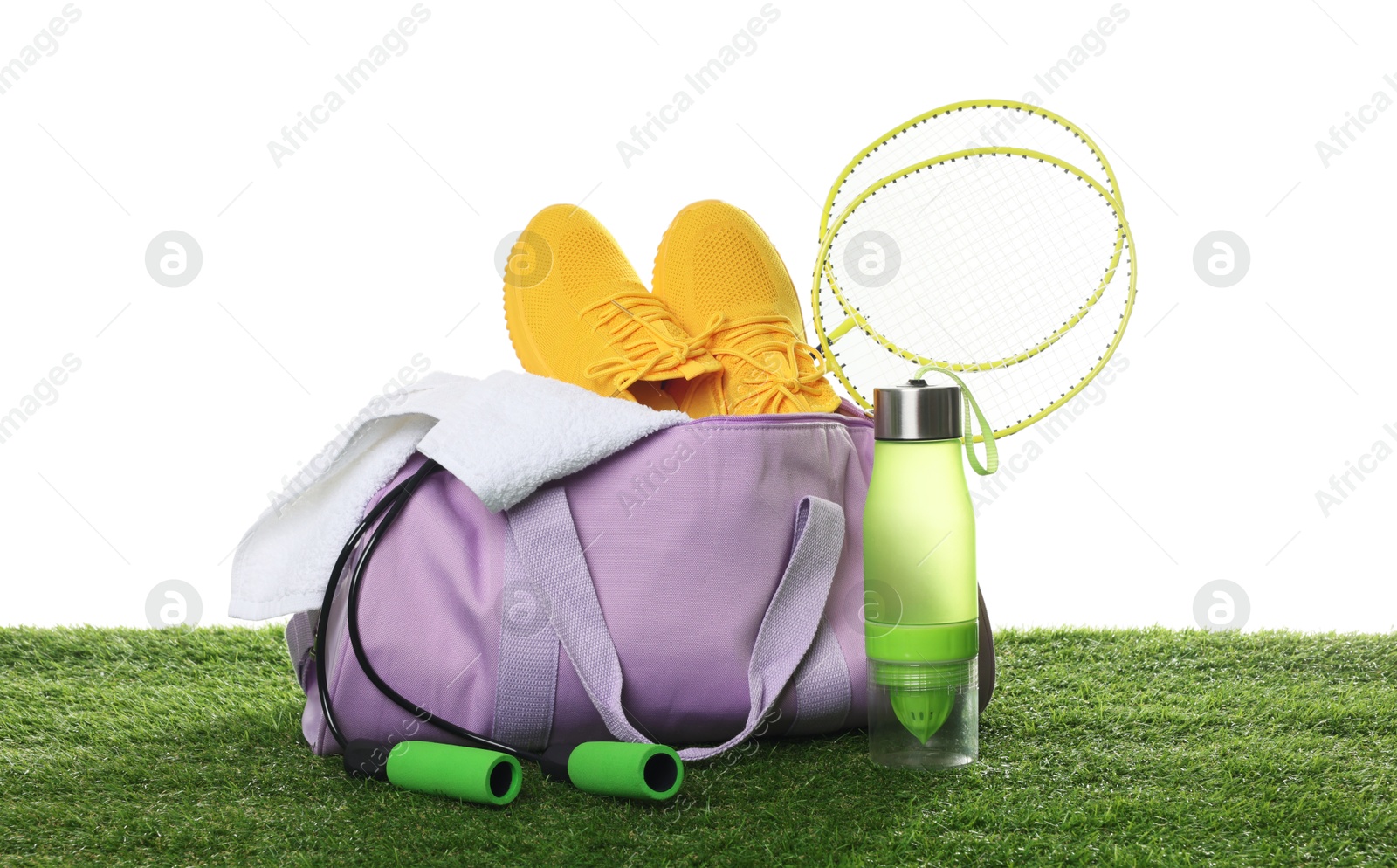 Photo of Stylish sports bag with badminton rackets, towel, skipping rope, bottle and sneakers on green grass against white background