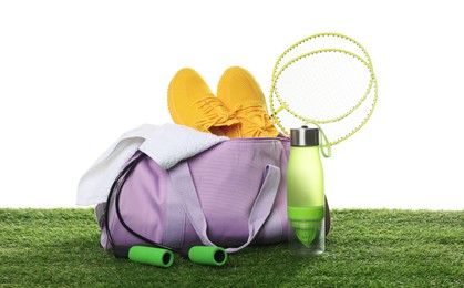Photo of Stylish sports bag with badminton rackets, towel, skipping rope, bottle and sneakers on green grass against white background