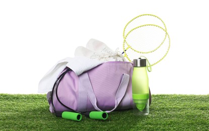 Photo of Stylish sports bag with badminton rackets, towel, skipping rope, bottle and sneakers on green grass against white background