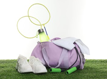 Photo of Stylish sports bag with badminton rackets, towel, skipping rope, bottle and sneakers on green grass against white background