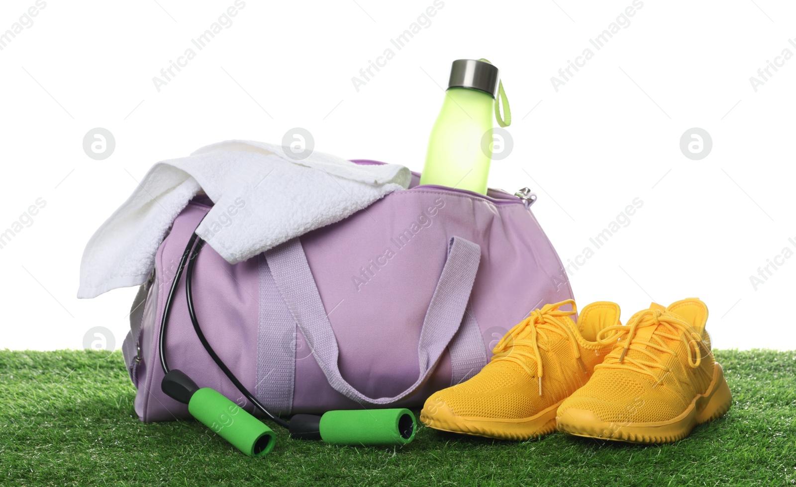 Photo of Stylish sports bag with bottle, towel, skipping rope and sneakers on green grass against white background