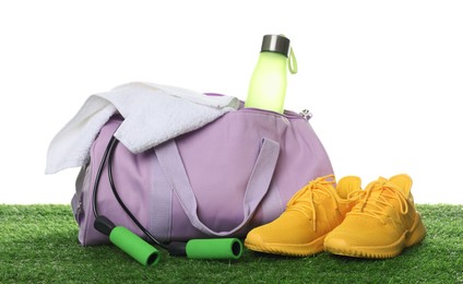 Photo of Stylish sports bag with bottle, towel, skipping rope and sneakers on green grass against white background
