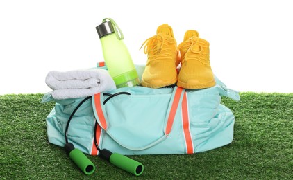 Photo of Stylish sports bag with skipping rope, towel, bottle and sneakers on green grass against white background
