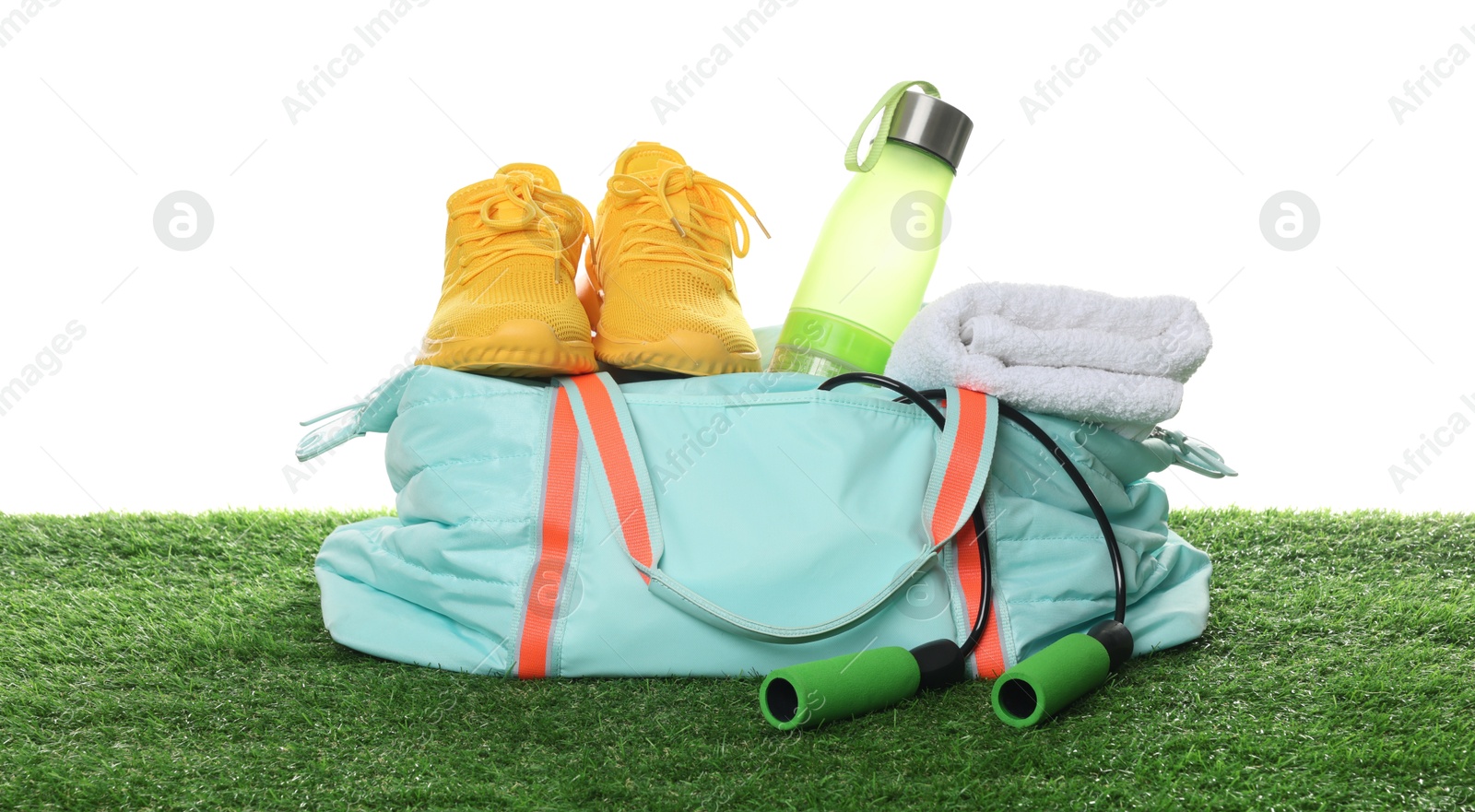 Photo of Stylish sports bag with skipping rope, towel, bottle and sneakers on green grass against white background