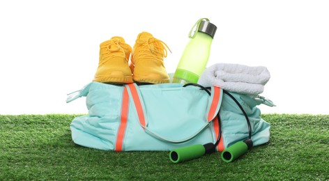 Photo of Stylish sports bag with skipping rope, towel, bottle and sneakers on green grass against white background