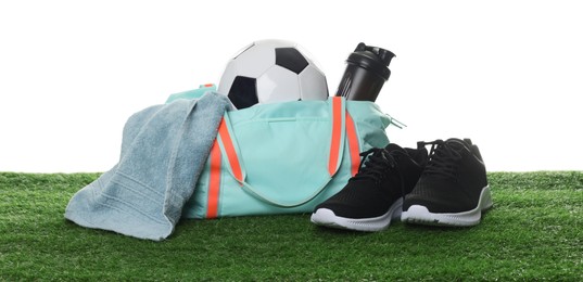 Photo of Stylish sports bag with soccer ball, towel, shaker cup and sneakers on green grass against white background