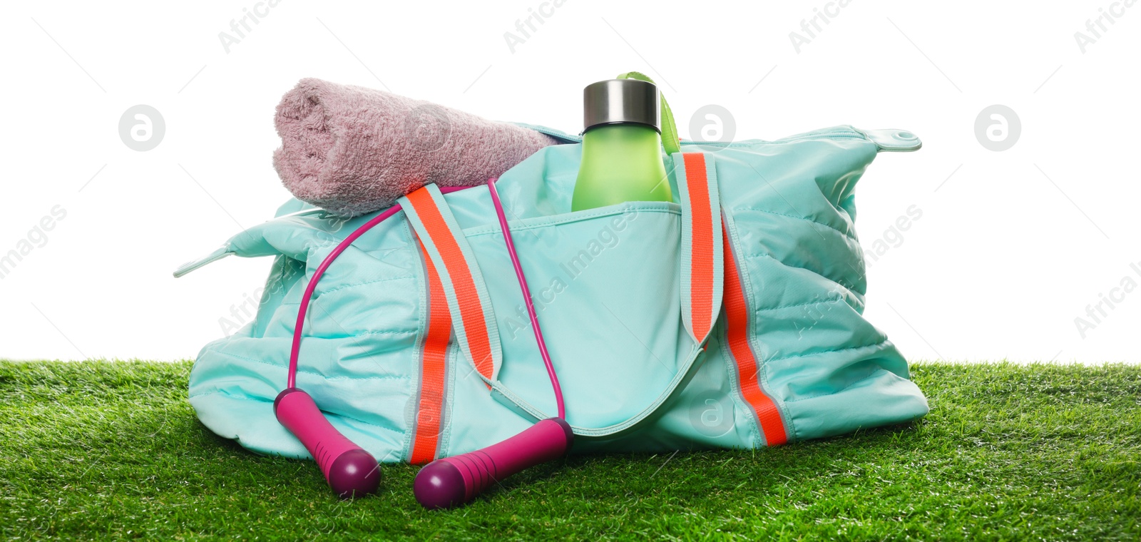 Photo of Stylish sports bag with skipping rope, towel and bottle on green grass against white background