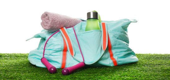 Photo of Stylish sports bag with skipping rope, towel and bottle on green grass against white background