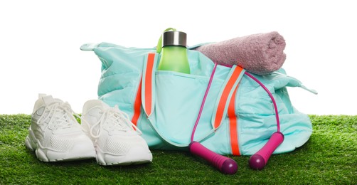 Photo of Stylish sports bag with skipping rope, towel, bottle and sneakers on green grass against white background