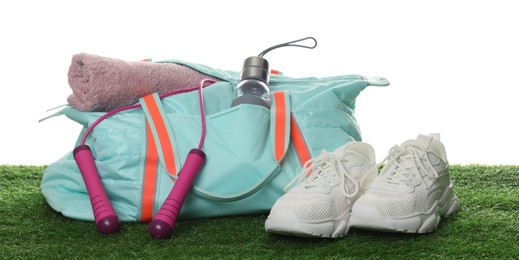 Photo of Stylish sports bag with skipping rope, towel, bottle and sneakers on green grass against white background
