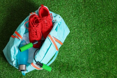 Photo of Stylish sports bag with skipping rope, bottle and sneakers on green grass, top view. Space for text