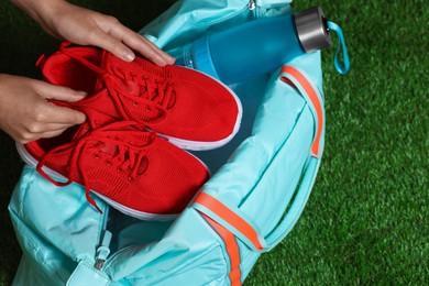Photo of Woman putting sneakers into stylish sports bag on green grass, top view