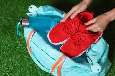 Photo of Woman putting sneakers into stylish sports bag on green grass, top view