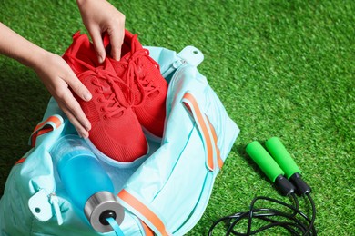 Photo of Woman putting sneakers into stylish sports bag on green grass, closeup