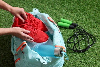 Photo of Woman putting sneakers into stylish sports bag on green grass, closeup
