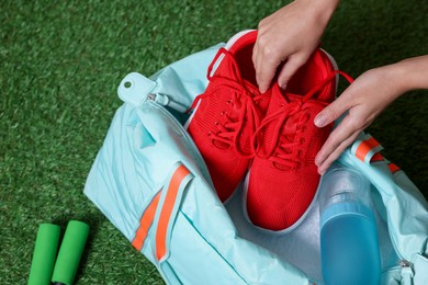 Photo of Woman putting sneakers into stylish sports bag on green grass, top view