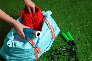 Photo of Woman putting sneakers and bottle into stylish sports bag on green grass, closeup