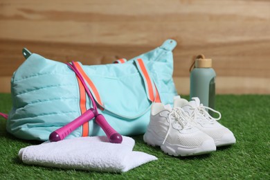 Photo of Stylish sports bag with skipping rope, towel and bottle on green grass against wooden background, closeup