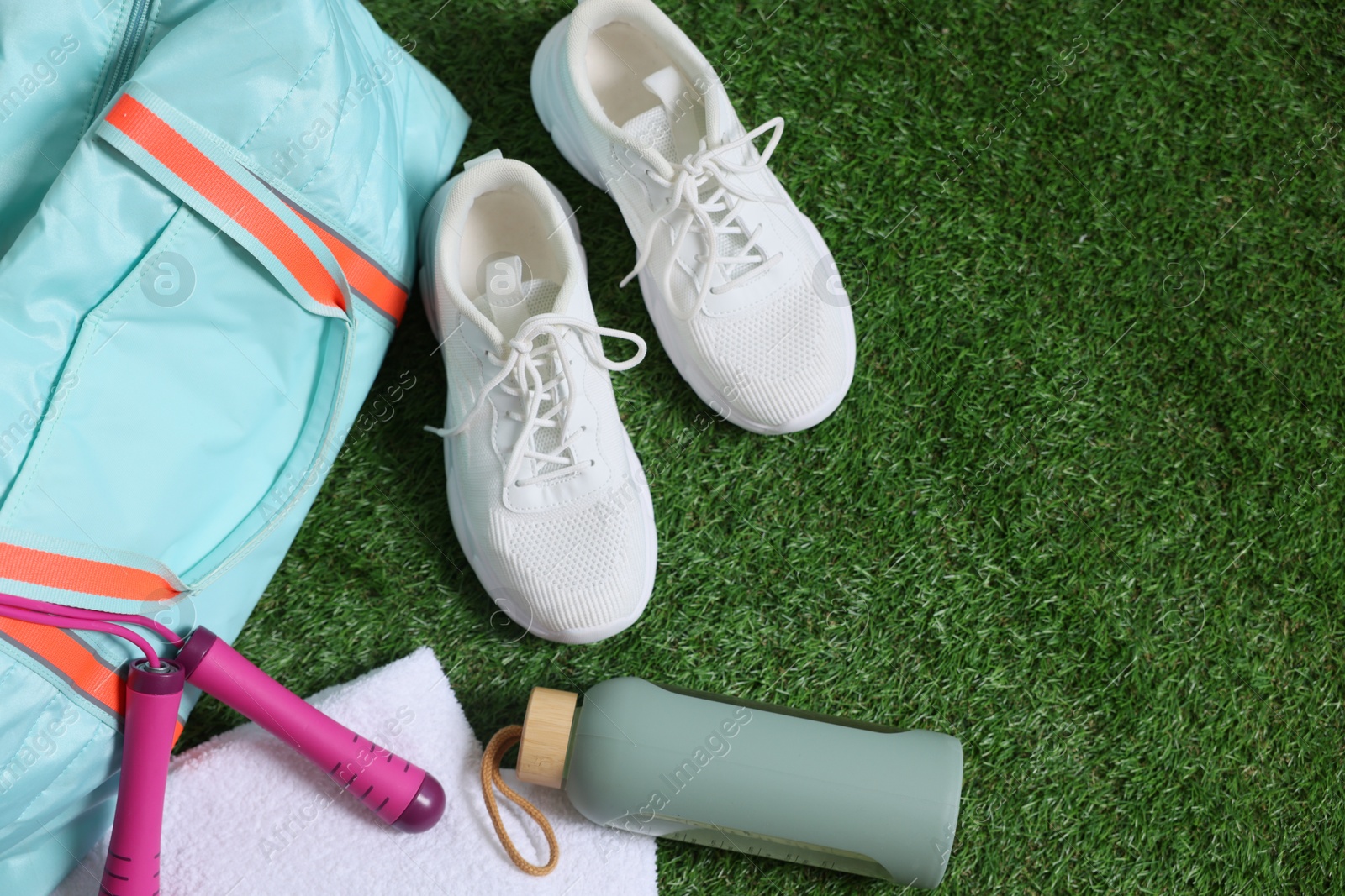 Photo of Stylish sports bag with skipping rope, towel, bottle and sneakers on green grass, flat lay. Space for text