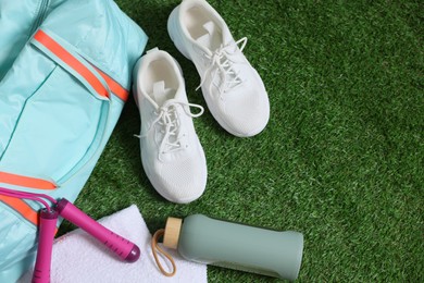 Photo of Stylish sports bag with skipping rope, towel, bottle and sneakers on green grass, flat lay. Space for text